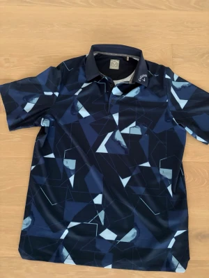 Mönstrad golfpiké från Callaway - Snygg mörkblå golfpiké från Callaway med geometriskt mönster i blått och vitt. Pikén har klassisk krage, knappar framtill och är tillverkad i ett lätt, snabbtorkande funktionsmaterial (Opti-Dri) som passar perfekt för sportiga aktiviteter.