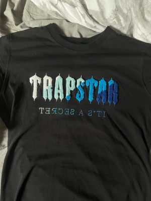 Trapstar svart t-shirt med brodyr - Säljer en svart t-shirt från Trapstar med broderad text i vitt, ljusblått och blått på bröstet. Texten 'TRAPSTAR' står i stora bokstäver och under står det 'IT'S A SECRET' i mindre stil. Klassisk passform och rund halsringning.