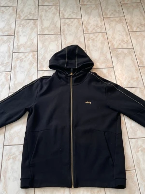 Svart zip från Boss - Snygg svart hoodie från BOSS med dragkedja framtill och diskret BOSS-logga på bröstet. Hoodien har huva med kontrastsömmar och två stora fickor framtill. Tillverkad i mjukt bomullsmaterial, perfekt för en avslappnad stil. storlek L men både M och L passar