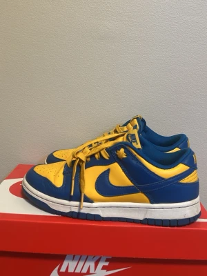 Nike Dunk Low blå/gula sneakers - Nike Dunk Low sneakers i blått och gult läder med vit sula och matchande gula skosnören. Klassisk låg siluett med perforerad tåbox och ikonisk Swoosh på sidan. Perfekta för dig som vill sticka ut med färg och stil.