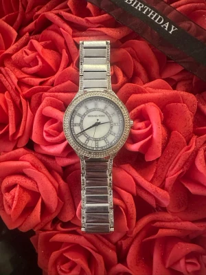 Silverklocka från Michael Kors - Stilren silverfärgad klocka från Michael Kors med länkarmband och glittrande stenar runt urtavlan. Urtavlan är vit med dekorativa detaljer och märkets logga. Perfekt accessoar för att lyfta din outfit.