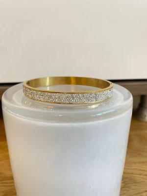 Guldigt armband med gnistrande stenar - Snyggt och stilrent armband i guldfärgad metall med en rad tätt sittande gnistrande stenar på ovansidan. Armbandet har en klassisk, rund form och ger en lyxig känsla till din outfit. Perfekt för dig som gillar bling och vill sticka ut lite extra.