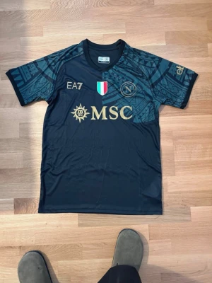 Napoli matchtröja EA7 svart - Säljer en svart Napoli matchtröja från EA7 med guldfärgade logotyper och MSC-tryck på framsidan. Tröjan har coola geometriska mönster på axlarna och är tillverkad i lätt och funktionellt material. Italienska flaggan och klubbens emblem finns på bröstet.