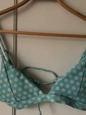 Ny Bikini i turkost med blommigt mönster - Supersöt bikini i turkos med små vita och gula blommor. Överdelen har smala axelband och snörning i ryggen för justerbar passform. Underdelen är klassisk med samma blommiga mönster. Perfekt för lata dagar på stranden!