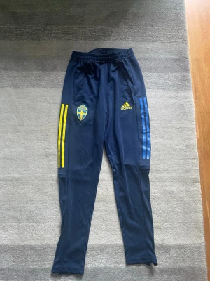 Blå adidas Sverige träningsbyxor - Mörkblå träningsbyxor från adidas med Sveriges landslagsemblem på ena benet och adidas-logga på andra. Byxorna har gula och blå ränder längs sidorna och är tillverkade i ett lätt, syntetiskt material. Perfekta för fotbollsträning eller chill kommande VM