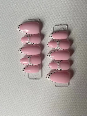 Rosa press-on naglar med prickig tipp - Snygga press-on naglar i ljusrosa med vita tippar dekorerade med svarta prickar. Naglarna har en mandelformad design och en glansig finish. Perfekt för dig som vill ha en lekfull och trendig look.