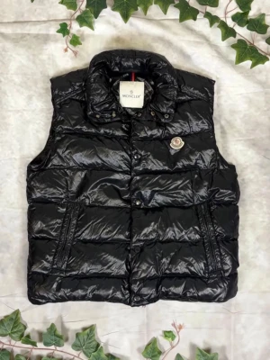 Svart dunväst från Moncler - Snygg svart dunväst från Moncler med glansig finish och hög krage. Västen har dragkedja och tryckknappar framtill, två fickor och klassisk Moncler-logga på bröstet. Perfekt för lager-på-lager och streetstyle.
