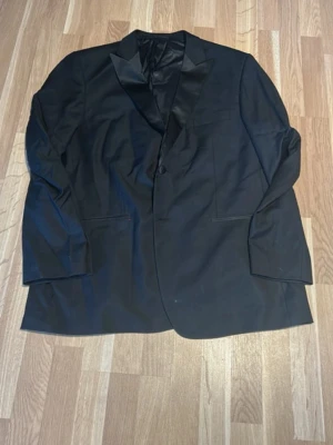 Svart kavaj från Dressmann, storlek 52 - Säljer en klassisk svart kavaj från Dressmann i Super 100's ull. Kavajen har ett snyggt satinliknande slag, enkelknäppning och två framfickor. Perfekt för dig som vill ha en stilren och tidlös look.
