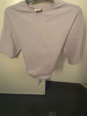Ljuslila topp med knyt i midjan - Trendig ljuslila topp med korta ärmar och rund halsringning. Toppen har en snygg knytning i midjan baktill som ger en cool och avslappnad look. Perfekt att matcha med jeans eller kjol. Materialet ser ut att vara mjukt bomullstyg.