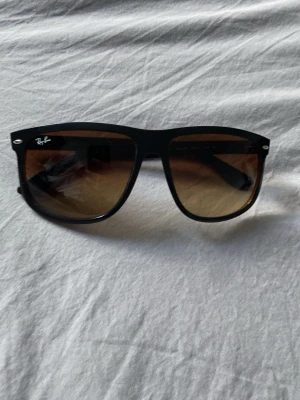 Ray-Ban solglasögon - Snygga svarta solglasögon från Ray-Ban med klassisk wayfarer-form och bruna tonade glas. Perfekta för soliga dagar och ger en cool vibe till din outfit. Diskreta silvriga detaljer vid bågarna och logga på linsen.