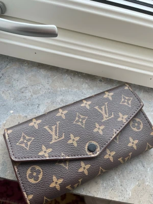  plånbok - Säljer en brun plånbok   med det klassiska monogrammönstret i beige. Plånboken har kuvertformad design och stängs med en knapp framtill. Tillverkad i tåligt canvasmaterial med exklusiv känsla och ikoniska detaljer .