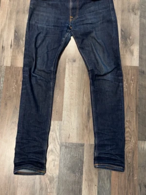 Mörkblå jeans från Nudie Jeans - Säljer ett par mörkblå jeans från Nudie Jeans med klassisk femficksdesign och snygga kontrastsömmar. Jeansen har smal passform och detaljerade bakfickor med Nudie Jeans signaturbroderi. Perfekt för dig som gillar stilrena och tidlösa jeans.