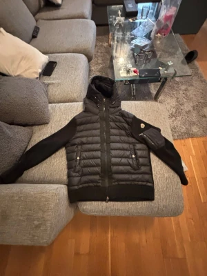 Svart pufferjacka från Moncler - Snygg svart pufferjacka från Moncler med quiltad front och rygg, samt släta ärmar. Jackan har huva, två dragkedjeförsedda fickor fram och en Moncler-logga på ärmen. Perfekt för kalla vinterdagar och riktigt stilren look.