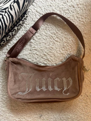 Brun axelväska Juicy Couture - Brun axelväska från Juicy Couture i mjukt velourmaterial. Väskan har ett glittrigt Juicy-tryck med stenar på framsidan och en bred axelrem med broderad logga. Perfekt för dig som gillar Y2K-stilen och vill ha något unikt.