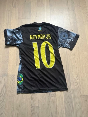 Brasil Neymar Jr. matchtröja svart - Säljer en svart Brasilien matchtröja från Nike med Neymar Jr. och nummer 10 i gult på ryggen. Framsidan har ett detaljerat grafiskt tryck med Kristusstatyn och brasilianska symboler. Tröjan har korta ärmar och rund hals. Perfekt för fotbollsfans!