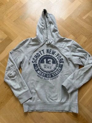 Grå hoodie från Schott NYC - Snygg grå vintage hoodie från Schott NYC med tryck i mörkblått framtill där det står 'Schott New York' och 'No West 33 Street'. Hoodien har dragsko i huvan och breda muddar vid ärmslut och nederkant. Perfekt för en avslappnad streetwear-look. Passar bäst för dig mellan 164-170, står S men passar XS