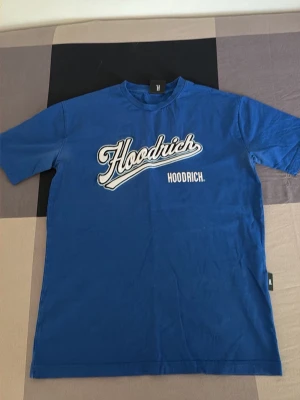 Blå Hoodrich t-shirt med tryck - Snygg blå t-shirt från Hoodrich med stort vitt tryck på bröstet och coolt tryck på ryggen. Klassisk passform med rund hals och korta ärmar. Perfekt för dig som gillar streetwear och vill sticka ut med en bold look.