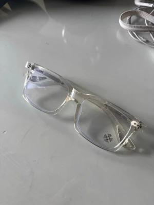 Chrome hearts glasögon - Säljer ett par chrome hearts glasögon med transparent plastbåge och rektangulär form. Glasen har en lätt blåtonad reflektion och det finns en liten dekorativ detalj på ena glaset. Perfekt accessoar för dig som gillar minimalistisk design.