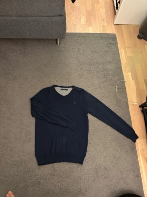 Mörkblå V-ringad tröja Tommy Hilfiger - Mörkblå V-ringad tröja från Tommy Hilfiger med diskret logga på bröstet. Tröjan har långa ärmar, ribbade muddar och är tillverkad i ett mjukt, tunt material som passar perfekt till lager-på-lager-stil.