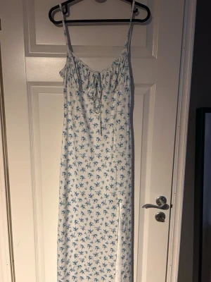 Vit långklänning med blå blommor - Långklänning med smala axelband och blått blommönster på vit botten. Klänningen har snörning vid bysten och en liten slits framtill.