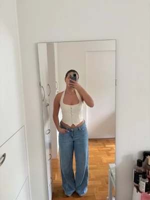 Cremevit korsettopp med knappar - Säljer en snygg vit korsettopp med halterneck och dekorativa knappar framtill. Toppen har en figurnära passform och är ärmlös, perfekt till jeans eller kjol. Materialet känns mjukt och stretchigt, troligen bomull eller polyester. Helt ny med prislappen kvar!