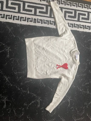 Vit stickad tröja från Ami Paris - Vit stickad tröja från Ami Paris med rund halsringning och ribbade muddar. På bröstet finns ett rött hjärta och bokstaven 'A' broderat. Tröjan har en klassisk passform och är perfekt för en stilren look. 