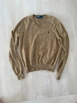 Beige v-ringad tröja från Polo Ralph Lauren - Klassisk beige v-ringad tröja från Polo Ralph Lauren med den ikoniska broderade loggan på bröstet. Tröjan har långa ärmar, ribbade muddar och är tillverkad i ett tunt, mjukt material som passar perfekt till lager på lager. Enkel och stilren design.