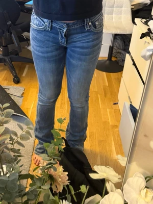 Blå bootcut jeans från Rock Revival - Säljer ett par blå bootcut jeans från Rock Revival med snygga slitningar och detaljerade bakfickor med broderi och nitar. Jeansen har låg midja och kontrastsömmar som ger en cool look. Perfekta för dig som gillar statement-jeans!