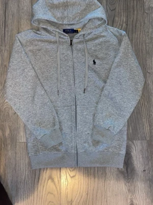 Grå zip hoodie från Polo Ralph Lauren - Snygg gråmelerad hoodie från Polo Ralph Lauren med dragkedja framtill och klassisk huva med dragsko. Ikoniska Polo-loggan broderad på bröstet. Tillverkad i mjukt bomullsmaterial och har ribbade muddar vid ärmar och nederkant. Perfekt för en chill och stilren look.