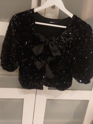 Svart paljettkofta med rosetter Gina Tricot - Glittrig svart topp från Gina Tricot. Två rosetter framtill. Modellen har korta puffärmar. Jag har använt den 1 gång och säljer för att den inte kommer till användning. Den är i storlek xs och är från Gina Tricot. 