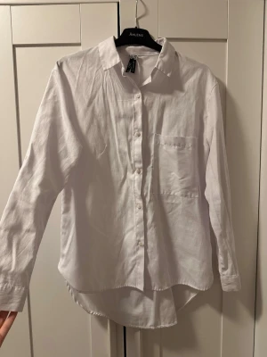 Vit linneskjorta  - Klassisk vit skjorta i linne med lång ärm, bröstficka och knappar framtill. Skjortan har en avslappnad passform och rundad nederkant, perfekt för en clean och enkel stil.