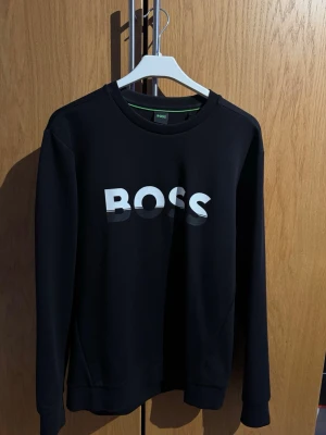 Svart sweatshirt från BOSS - Svart sweatshirt från BOSS med stor vit logga på bröstet. Snygg och enkel design som passar perfekt till jeans eller joggers. Äkta produkt och köpt från Boozt. Kan gå ned i pris vid snabb affär!
