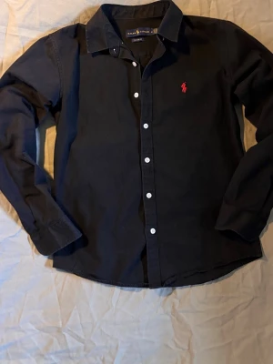 Svart skjorta från Ralph Lauren - Klassisk svart skjorta från Ralph Lauren med vit knappstängning och röd broderad logga på bröstet. Skjortan har lång ärm, klassisk krage och är tillverkad i bomull för en bekväm och stilren look.