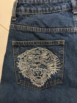 Blå Ed Hardy jenas med glittrig tiger på fickan - Snygga blå Ed Hardy jeans med en glittrig tiger i silver på bakfickan. Jeansen har klassisk femficksdesign och är gjorda i denim. Perfekta för dig som vill sticka ut med coola detaljer och streetstyle-vibe.