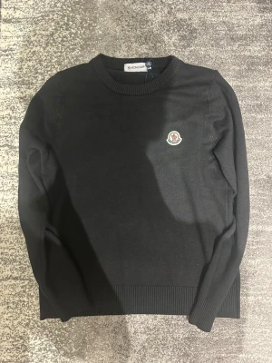 Svart stickad tröja från Moncler - Snygg svart stickad tröja från Moncler med rund halsringning och Moncler-logga på bröstet. Tröjan har långa ärmar och ribbade muddar vid ärmslut och nederkant. Perfekt för dig som gillar stilrena plagg med exklusiv känsla. OBS! Tröjan är mindre där den passar folk som är ca 170cm alltså XS/S, mittemellan 