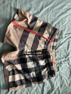Burberry rutiga badbyxor xxl - Säljer ett par klassiska badbyxor från Burberry i ikoniskt beige, svart, vitt och rött rutmönster. Modellen har resår i midjan med snörning och innerbyxa i mesh. Perfekta för stranden eller poolen, riktigt snygg och tidlös design.