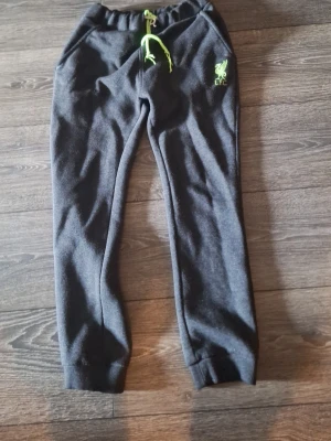 Svarta LFC mjukisbyxor med limegrönt tryck - Mjukisbyxor i svart med limegröna detaljer från Liverpool FC. Byxorna har resår i midjan med dragsko och ribbade muddar vid bensluten. LFC-logga i limegrönt på ena benet. Perfekta för chill eller träning.