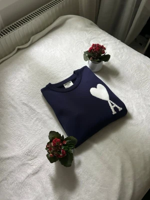 Mörkblå Ami Paris tröja med hjärta - Säljer en mörkblå stickad tröja från Ami Paris med ett vitt hjärta och bokstaven A på bröstet. Tröjan har rund halsringning och ribbade muddar. Perfekt för dig som gillar stilrena och trendiga plagg med en twist.