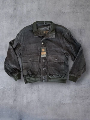 Mörkbrun Läderjacka i mockastil - Vintage Style (Oversize) - Säljer en mörkbrun jacka i äkta läder. Utsidan har en mockaliknande finish och insidan är fodrad med polyester. Jackan är oanvänd och har kvar originaltags, men har förvarats i en flyttkartong under en period och kan därför behöva vädras/tvättas innan användning.  Detaljer: • Storlek: XL (något oversize) • Färg: Mörkbrun • 2 ytterfickor med lock • 2 innerfickor, varav en av de är med dragkedja • Ribbad krage, muddar och nederkant  Fler bilder finns att skicka. Skriv om du har några frågor!