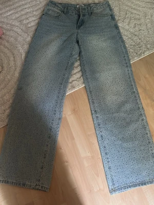 Ljusblå vida jeans med strassdetaljer - Säljer ett par ljusblå jeans med raka, vida ben och glittrande strassdetaljer över hela tyget. Jeansen har klassisk femficksmodell och stängs med knapp och dragkedja. Perfekta för dig som vill sticka ut lite extra!