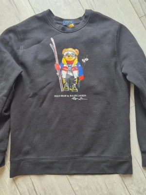 Svart sweatshirt Polo Bear Ralph Lauren - Cool svart sweatshirt från Ralph Lauren med Polo Bear-tryck på bröstet. Björnen har skidutrustning och färgglada kläder. Tröjan har rund halsringning och långa ärmar. Perfekt för dig som gillar streetstyle och klassiska märken.