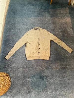 Grå cardigan från Polo Ralph Lauren - Snygg ljusgrå cardigan från Polo Ralph Lauren med mörkblå broderad logga på bröstet. Tröjan har V-ringning, knäppning med svarta knappar och ribbade muddar. Perfekt att slänga över en t-shirt för en clean look.