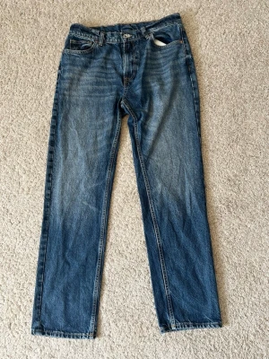 Blå raka jeans i klassisk modell - Säljer ett par blå jeans med rak passform och klassisk femficksdesign. Jeansen har en snygg tvätt med lätt slitning och är tillverkade i denim. Perfekta för dig som gillar en tidlös och enkel stil.