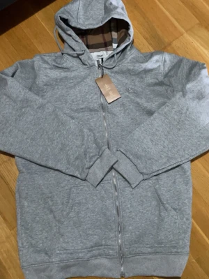 Grå zip hoodie från Burberry - Snygg grå zip hoodie från Burberry med klassiskt rutigt foder i huvan och dragkedja framtill. Tillverkad i mjukt bomullsmaterial och har två fickor fram. Perfekt för en chill och stilren look. Komm med bud vid snabb affär 
