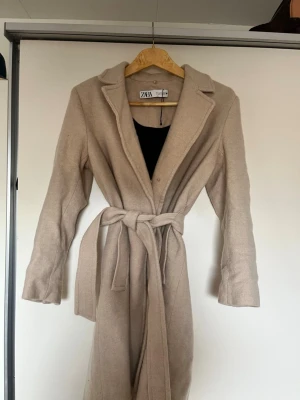 Beige kappa med knytskärp från Zara - Stilren beige kappa från Zara med klassisk krage och knytskärp i midjan. Kappan har en rak passform och är tillverkad i ett mjukt tyg som ger en elegant look. Perfekt för dig som vill ha en enkel men snygg ytterjacka till hösten eller våren. Man kan lägga till krage om man vill använd 3-4 gånger i vintras 