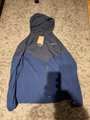 Nike blå träningsset med huva - Snyggt träningsset från Nike med en tvåfärgad blå och mörkblå jacka med huva och dragkedja samt matchande blå träningsbyxor med resår i midjan och Nike-logga. Perfekt för löpning eller gym. Lätt material som andas och ger bra rörelsefrihet.