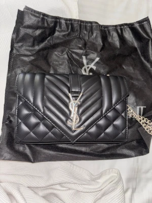 Svart YSL quiltad handväska - Säljer en svart quiltad handväska från Yves Saint Laurent med silverfärgad kedjerem och ikonisk YSL-logga framtill. Väskan har ett elegant kuvertformat lock och är tillverkad i slätt läder. Dustbag medföljer samt en korthållare matchande med väskan vid snabbt köp☺️ Kom gärna med prisförslag ifall ni är intresserade!