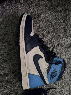 Nike Air Jordan 1 High obsidian  - Säljer ett par Nike Air Jordan 1 High sneakers i blått, vitt och marinblått. Skorna har klassisk hög siluett, svart swoosh och detaljer i läder. Perfekta för dig som gillar streetwear och vill sticka ut med en ikonisk modell.