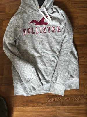 Grå Hollister hoodie med logga - Snygg gråmelerad hoodie från Hollister med stor vinröd logga och text på bröstet. Tröjan har huva med vita snören och en klassisk magficka. Perfekt för en avslappnad och trendig stil. Tillverkad i mjukt och bekvämt material.