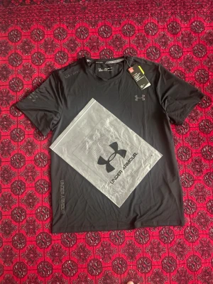 Svart Under Armour t-shirt - Snygg svart t-shirt från Under Armour med färgglad logga på bröstet och tryckt text på axeln och sidan. Perfekt för träning eller chill. Tillverkad i ett lätt och snabbtorkande material som känns skönt mot huden. Klassisk passform och rund hals.